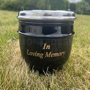 Puede incluir: Una urna funeraria de cerámica negra con acabado brillante y tapa de plata metálica. La parte frontal de la urna presenta el texto dorado In Loving Memory, centrado en una fuente serif. El recipiente tiene un cuerpo redondeado y una superficie lisa.