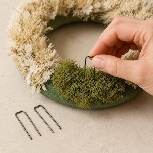 May include: A circular wreath base featuring a section of dried beige grasses and small white flowers alongside a patch of vibrant green moss. Three dark metal U-shaped floral pins are included, with one being inserted into the moss.