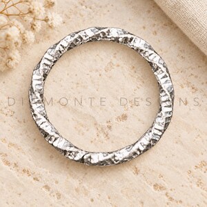 May include: A circular silver-toned metal ring featuring a highly textured, hammered surface with deep indentations and a reflective finish. The text "DIAMONTE DESIGNS" is clearly visible across the centre of the ring in a clean, sans-serif font.