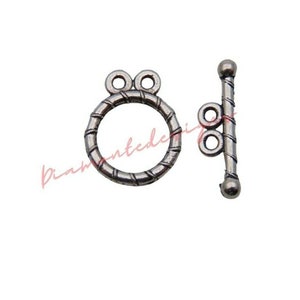 May include: Silver tone metal toggle clasp with a twisted design. The clasp has a round ring with two loops and a straight bar with a round end.