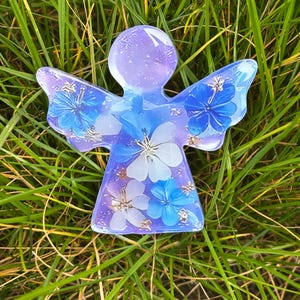 Pode incluir: Uma estatueta de anjo de resina com flores azuis e brancas embutidas e flocos dourados. O anjo tem um corpo lavanda e asas azuis, repousando sobre grama verde. As asas são translúcidas e as flores são visíveis.
