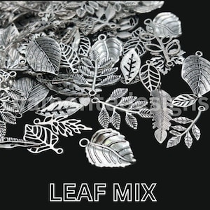 May include: A collection of silver-toned metal charms shaped like various leaves. Each charm features intricate vein detailing and a small loop at the top for attachment. The assortment includes diverse foliage styles, from broad rounded leaves to slender fern-like fronds, all displayed together. The text LEAF MIX is printed at the bottom.
