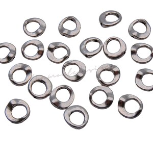 May include: A collection of small, silver-toned metal rings with an irregular, organic shape. The rings are scattered across a white surface, each with a slightly uneven texture and a hole in the center. Ideal for jewelry making.