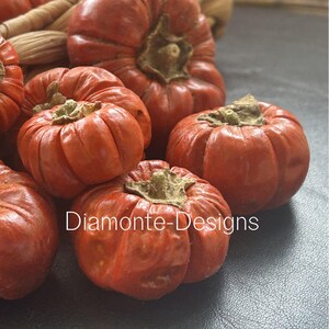 May include: A cluster of small, round, orange-red pumpkins with deep vertical ridges and brown, dried stems. The text Diamonte-Designs is printed in white across the center of the pumpkins, which are arranged in a group with a woven texture visible.