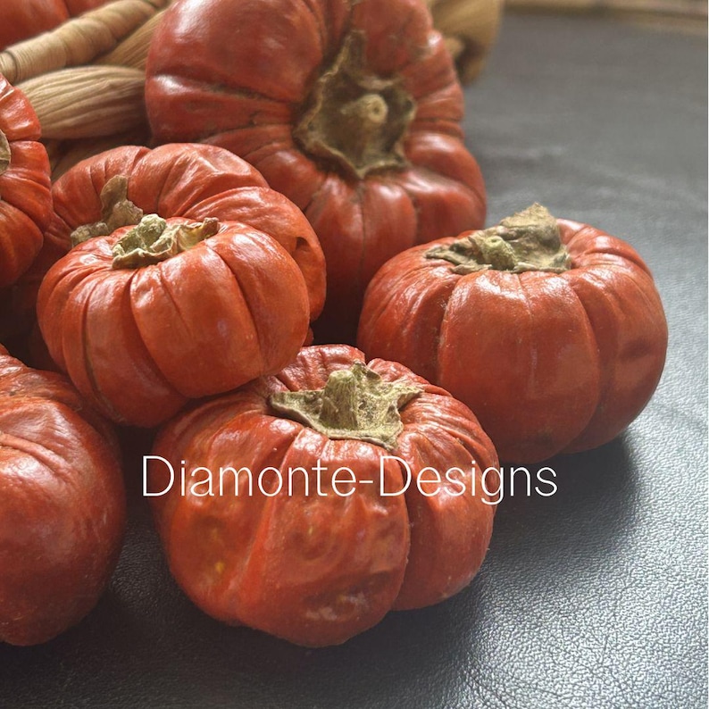 May include: A cluster of small, round, orange-red pumpkins with deep vertical ridges and brown, dried stems. The text Diamonte-Designs is printed in white across the center of the pumpkins, which are arranged in a group with a woven texture visible.
