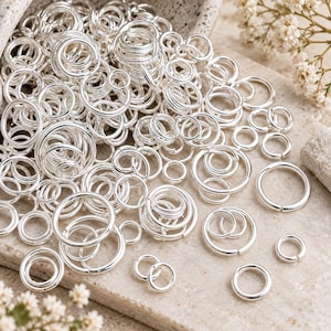 May include: A large collection of shiny silver-toned metal jump rings in various sizes. The circular rings feature a smooth, polished finish and a small split opening, commonly used for jewellery making and connecting various craft components.