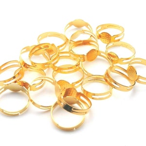 Puede incluir: Una colección de bases de anillos ajustables de color dorado. Cada anillo presenta una base plana y redonda y un diseño de banda dividida. Los anillos están dispersos sobre una superficie blanca, mostrando su acabado metálico brillante.