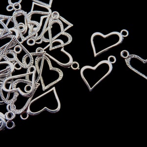 May include: Silver-colored heart-shaped charms with a small loop at the top for stringing. The charms are scattered on a black background. Some hearts are solid, while others have open designs.