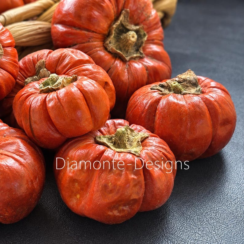 May include: A collection of vibrant orange, deeply ribbed pumpkins with textured brown stems, arranged in a cluster. The text "Diamonte-Designs" is printed in white across the lower center of the image, overlaying the pumpkins on a dark surface.