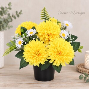 Könnte beinhalten: Ein Blumenarrangement in einem schwarzen Topf mit leuchtend gelben, dahlienartigen Blüten, kleinen weißen Gänseblümchen mit gelber Mitte und grünem Blattwerk. Der Text DiamonteDesigns ist in der oberen rechten Ecke des Bildes zu sehen.