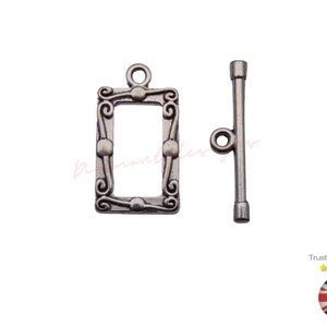 Op de afbeelding: Zilverkleurige metalen schuifgesp met een decoratief frame-ontwerp. De gesp bestaat uit twee delen: een rechthoekig frame met een decoratieve rand en een rechte stang met een lus aan het ene uiteinde.