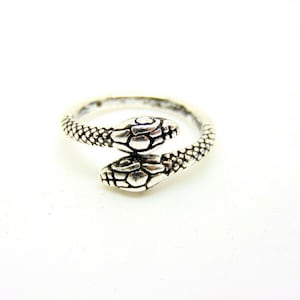 May include: A silver ring featuring two snake heads facing each other. The ring is crafted from textured metal and has a simple design.