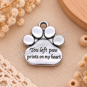 Op de afbeelding: Een zilverkleurige metalen bedel in de vorm van een dierenpootafdruk. De bedel heeft een klein oogje aan de bovenkant voor bevestiging en is gegraveerd met de zwarte tekst, You left paw prints on my heart, in het midden van de poot.