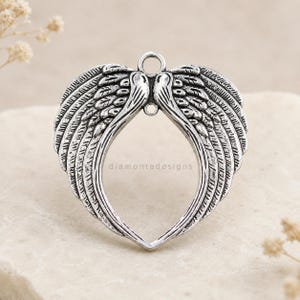 May include: A silver-toned metal pendant shaped like a pair of open angel wings forming a heart-like frame. The piece features detailed feather textures and includes two small circular loops at the top for attachment. The text diamontedesigns is visible.