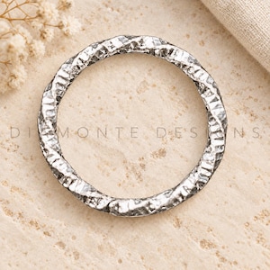 May include: A circular silver-toned metal ring featuring a highly textured, hammered surface with deep indentations and a reflective finish. The text "DIAMONTE DESIGNS" is clearly visible across the centre of the ring in a clean, sans-serif font.