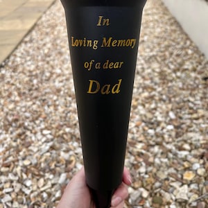 Puede incluir: Un jarrón de flores conmemorativo negro con una parte superior acampanada y una base puntiaguda. El texto dorado dice "In Loving Memory of a dear Dad". Una persona sostiene el jarrón.