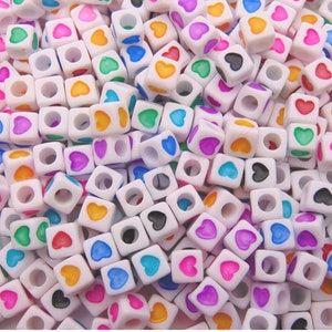 May include: A collection of white cube beads, each decorated with a heart in a range of colours: red, pink, yellow, green, blue, purple, and black. The beads have a hole through the centre, and are scattered together.
