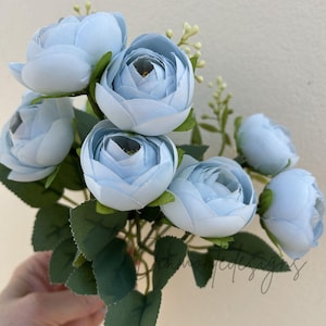 May include: A bunch of pale blue artificial flowers with green foliage. The flowers are fully open, displaying multiple petals. The arrangement includes small, white buds and is set against a plain background.