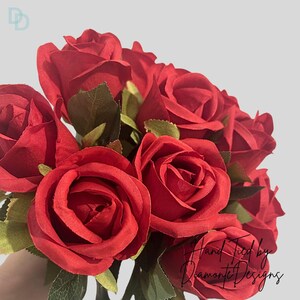 May include: A bouquet of deep red roses with green foliage. The roses are fully open, displaying their soft petals. The text "Hand Tied by Diamonte Designs" is visible in the lower right corner.
