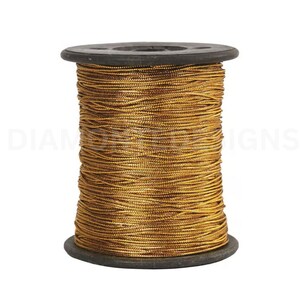 May include: A spool of metallic gold thread wrapped around a dark gray cylindrical bobbin. The thread has a reflective, shimmering texture and is wound tightly in horizontal layers. The text DIAMOND DESIGNS is visible in the background.