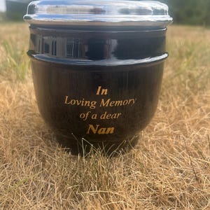 Może przedstawiać: Czarna ceramiczna urna z polerowaną srebrną metalową pokrywką. Z przodu urny znajduje się wygrawerowany złoty napis o treści: In Loving Memory of a dear Nan. Naczynie ma zaokrąglony korpus i lekko zwężającą się podstawę u dołu.