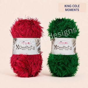May include: Two balls of King Cole Moments yarn, one in deep red and one in forest green. Each ball features a white paper label with the text King Cole Moments and a small crown logo, showcasing a fuzzy, eyelash-style texture throughout.