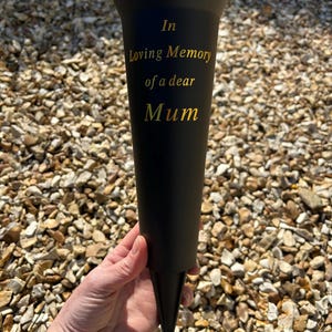 May include: Black memorial cone with gold lettering that reads "In Loving Memory of a dear Mum." The cone has a pointed base and is set against a backdrop of small, light-colored stones.