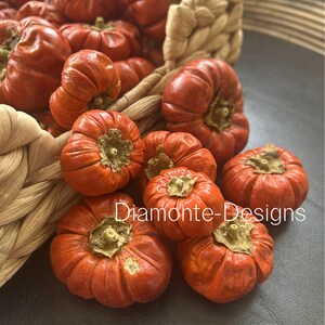 May include: A collection of small, vibrant orange, ribbed decorative pumpkins spilling out of a woven light tan basket. The pumpkins feature prominent brown stems and textured, rounded surfaces. The text Diamonte-Designs is visible.