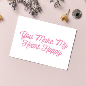 May include: White card with pink cursive text that reads "You Make My Heart Happy".