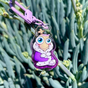 May include: A clear acrylic keychain featuring a cartoon king character with a crown, blue eyes, and a purple robe. The keychain has a purple clasp and is hanging against a backdrop of green succulent plants.