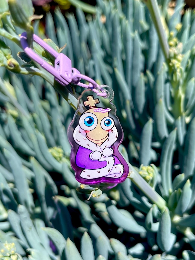 May include: A clear acrylic keychain featuring a cartoon king character with a crown, blue eyes, and a purple robe. The keychain has a purple clasp and is hanging against a backdrop of green succulent plants.