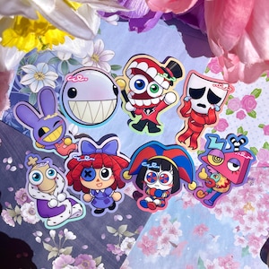 May include: A collection of colorful cartoon character stickers. The stickers feature various designs, including a smiling white ball with teeth, a red character in a top hat, and a sad face. The stickers are arranged on a floral background.