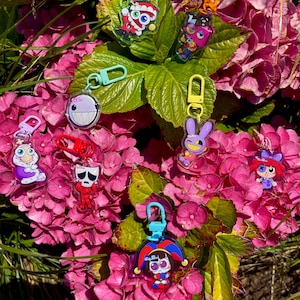 May include: A collection of colorful acrylic keychains featuring various cartoon characters. The keychains have different shapes and designs, including a jester, a bunny, and other whimsical figures. Each keychain has a metal clasp for attachment.