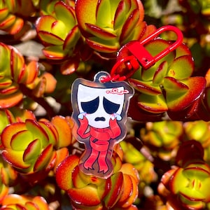 May include: A clear acrylic keychain featuring a cartoon character with a sad expression, wearing a red scarf and outfit. The keychain is attached to a red clasp. The background is a close-up of a succulent plant with green, orange, and red leaves.
