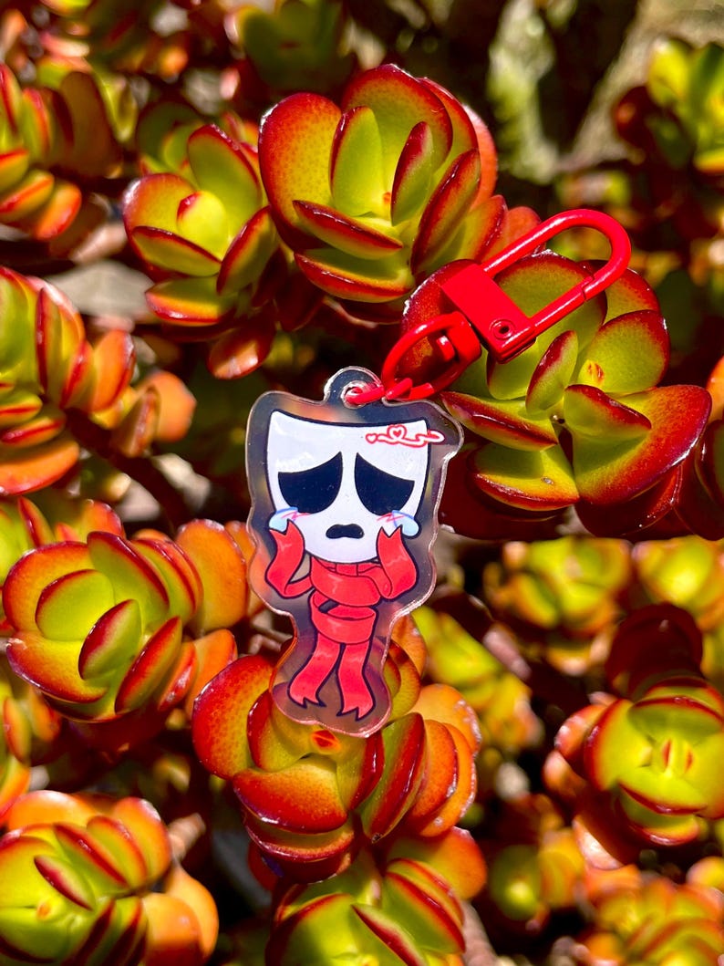 May include: A clear acrylic keychain featuring a cartoon character with a sad expression, wearing a red scarf and outfit. The keychain is attached to a red clasp. The background is a close-up of a succulent plant with green, orange, and red leaves.