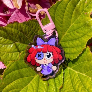 May include: A clear acrylic keychain featuring a cartoon doll with red hair, a purple dress, and a blue bow. The keychain has a pink clasp and is set against a backdrop of green leaves and pink flowers.