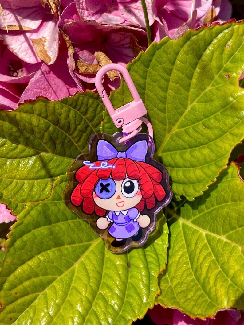 May include: A clear acrylic keychain featuring a cartoon doll with red hair, a purple dress, and a blue bow. The keychain has a pink clasp and is set against a backdrop of green leaves and pink flowers.