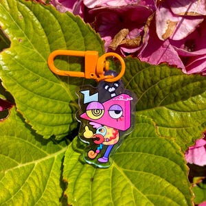 May include: A colorful acrylic keychain featuring a cartoon character with a pink, yellow, and blue design. The keychain has an orange clasp and ring. The background includes green leaves and pink flowers.