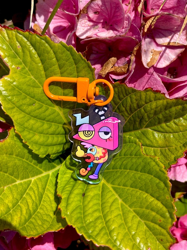 May include: A colorful acrylic keychain featuring a cartoon character with a pink, yellow, and blue design. The keychain has an orange clasp and ring. The background includes green leaves and pink flowers.