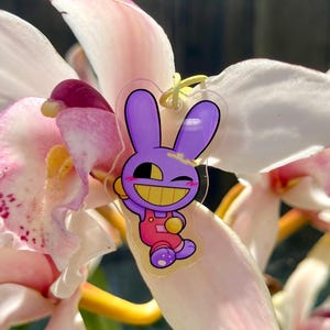 May include: A clear acrylic charm featuring a cartoon purple bunny with a yellow face and pink overalls. The charm is hanging on a yellow string, set against a backdrop of pink and white orchid petals.