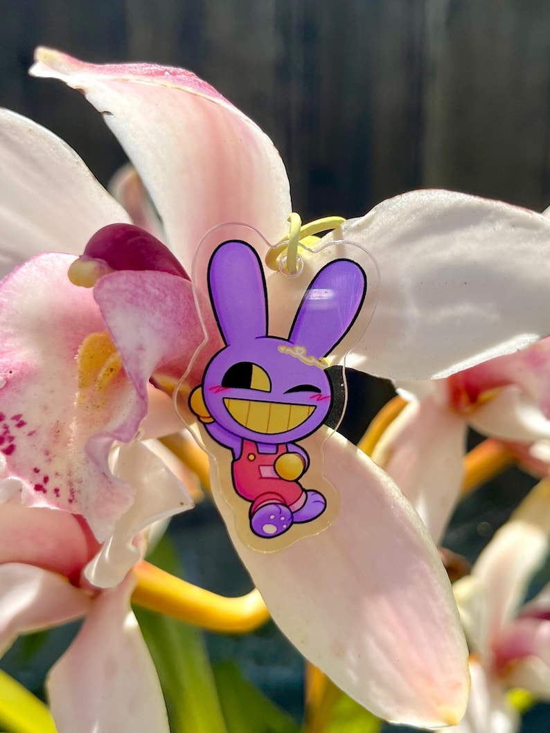 May include: A clear acrylic charm featuring a cartoon purple bunny with a yellow face and pink overalls. The charm is hanging on a yellow string, set against a backdrop of pink and white orchid petals.