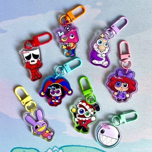 May include: A collection of acrylic keychains featuring colorful cartoon characters with various designs. Each keychain has a different character with unique features, attached to a metal clasp in various colors like red, orange, and green.