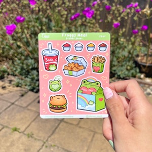 May include: A sticker sheet with a pink background featuring a froggy meal theme. The sheet includes stickers of a frog holding a soda, a frog holding a box of chicken nuggets, a frog holding a box of french fries, a frog holding a burger, and a frog holding a box of fries with a rainbow on top. The text "Froggy Meal" and "Sticker Sheet" are printed on the sheet.