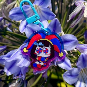 May include: A colorful keychain featuring a cartoon jester character with a blue clasp. The jester has a red and blue hat, and is set against a backdrop of purple flowers.