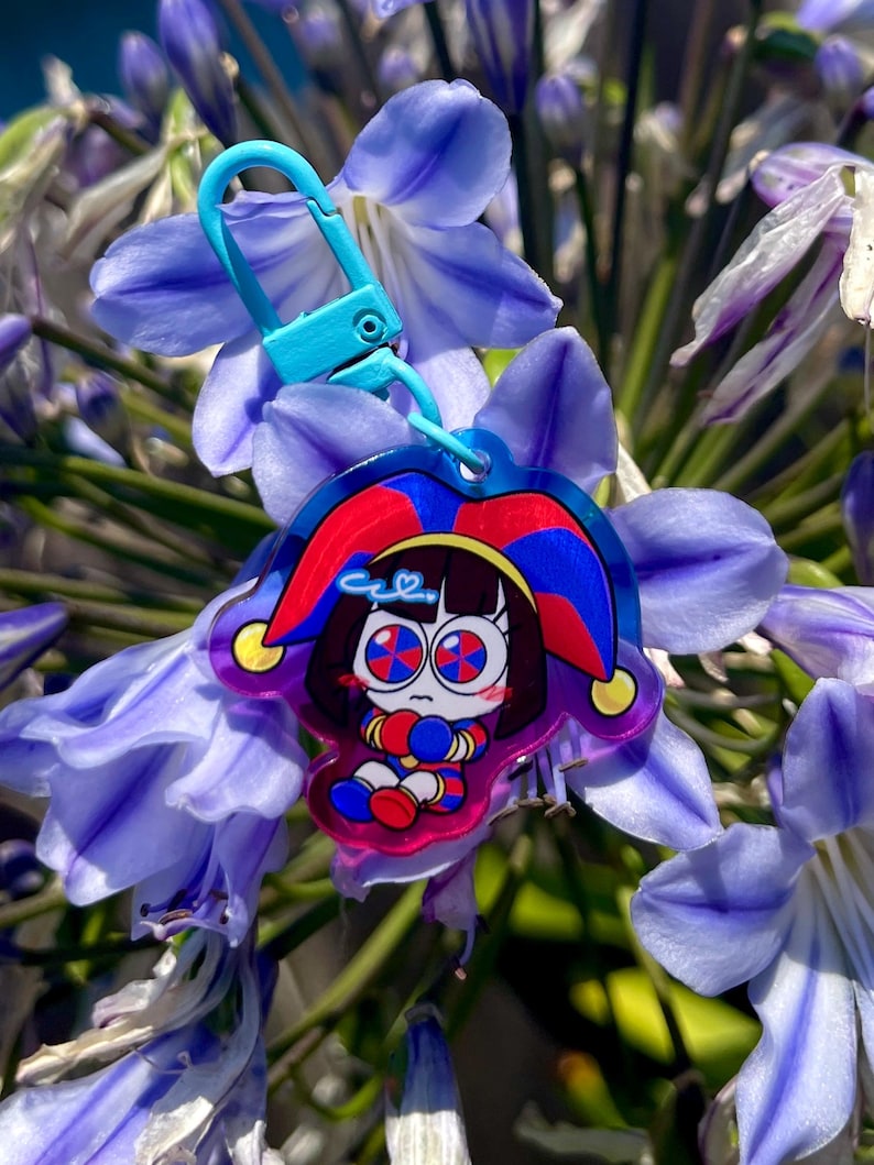 May include: A colorful keychain featuring a cartoon jester character with a blue clasp. The jester has a red and blue hat, and is set against a backdrop of purple flowers.