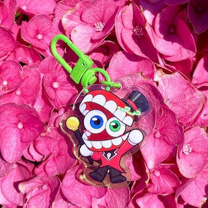 May include: A cartoon character keychain with a red top hat and suit, holding a yellow-topped cane. The keychain has a green clasp and is set against a backdrop of pink hydrangea flowers.