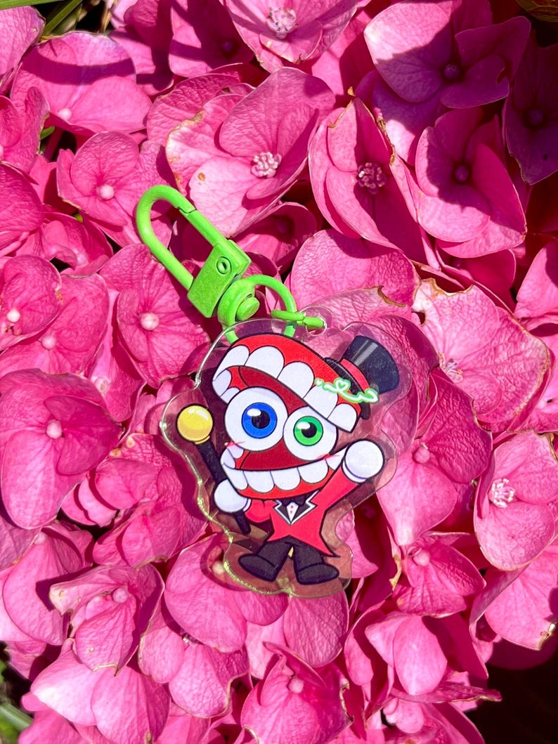 May include: A cartoon character keychain with a red top hat and suit, holding a yellow-topped cane. The keychain has a green clasp and is set against a backdrop of pink hydrangea flowers.
