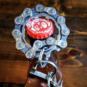Può includere: Un apribottiglie argentato a forma di catena da bicicletta con un portachiavi in ​​cuoio marrone attaccato. L'apribottiglie sta aprendo un tappo di bottiglia rosso con le parole "New Belgium Brewing" su di esso.
