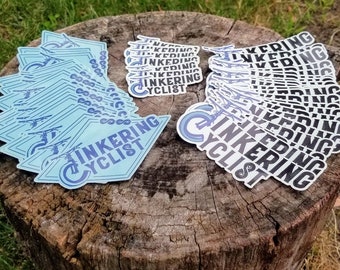 Puede incluir: Dos pilas de pegatinas con el texto "Tinkering Cyclist" en azul y blanco sobre un fondo azul claro. Las pegatinas están apiladas sobre un tocón de árbol.