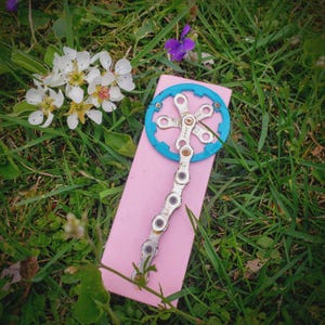 Op de afbeelding: Een handgemaakte flesopener gemaakt van een fietsketting en een blauw tandwiel, op een roze rechthoek. Witte bloemen en groen gras vormen de achtergrond. Een uniek, upcycled item.